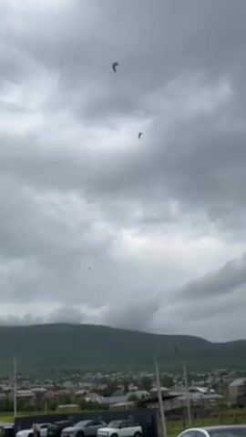 The video shows a view of a town and a mountain range on a cloudy day. There are several cars parked in the foreground. In the sky, there are a few birds flying. The scene is overcast with dark gray clouds.