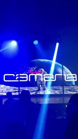 The video shows a group of people enjoying themselves at a club or event with a strong blue lighting effect. A screen in the background displays the word "Camara" in white, neon-style letters, with an image of an astronaut standing on a lunar-like surface under a large planet. Blue spotlights shine across the room, illuminating the dancing crowd. The overall atmosphere is energetic and festive.