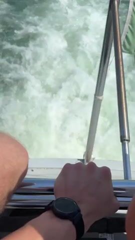 From the stern of a moving boat, the camera captures the wake churning white against the darker green water. A metal railing and a flag secured to it are visible on the right side of the frame. A person’s arm and hand holding the railing are visible at the bottom left corner. In the distance, land, buildings, and other boats can be seen under a clear blue sky.