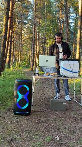 A man wearing headphones and a dark jacket stands behind a portable table in a forest. On the table, there is a laptop, a bottle of liquid, and a white container with blue straps. In front of the table, to the left, is a large, black JBL speaker with colorful lights. To the right of the table and on the ground is a grey battery. The man appears to be operating the laptop and the speaker. The setting is an outdoor environment with tall trees and green foliage.
