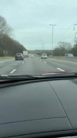 The video shows a driver's perspective from inside a red car moving along a multi-lane highway. Other cars and trucks are visible on the highway, moving in the same direction. The sky is overcast and gray. The road surface appears wet. Trees line the sides of the highway. Overhead, power lines run parallel to the road. The dashboard of the car is visible in the foreground, with the windshield and wipers above.