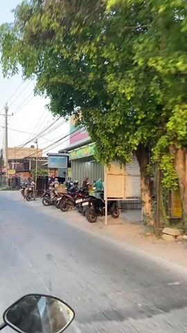 The video shows a street scene with several motorcycles parked outside a store. The store appears to be a small business, possibly a convenience store or a fruit stand, with a sign that reads "Laskar Buah." There are a few people visible in the video, some of whom are interacting with each other. One person is holding a child. The street is paved and there are buildings on either side of the road. Overhead wires are visible, suggesting an urban or suburban setting. The time of day appears to be late afternoon or early evening, based on the lighting.