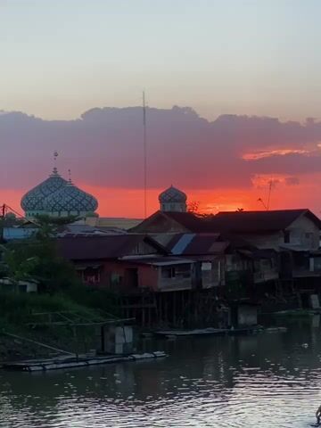 The video shows a sunset over a village with houses built on stilts next to a river. A mosque with a dome is visible in the background. Several children are swimming in the river, and an adult is sitting on a raft. The sky is filled with orange and pink clouds. The water is murky and reflects the sky.
