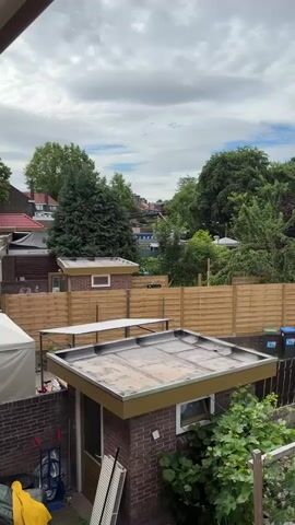 This media file captures an outdoor scene of a backyard in Heerlen, Netherlands, under a cloudy sky. The foreground features the roof of a small brick shed with a greenish-gold trim, suggesting a residential area. To the left, a white canvas tent or shelter is partially visible, along with a brick wall adorned with some gardening tools, including a red-handled garden cart and a yellow garment draped over a dark tarp. A green watering can is also present.

Behind the shed, a light brown wooden fence separates the properties. Beyond the fence, there are more brick structures, likely other sheds or small buildings, some with peaked roofs and windows. A tall evergreen tree stands prominently in the midground, contributing to the lush greenery that fills the background, interspersed with deciduous trees and the rooftops of houses further in the distance. The clouds are heavy and diffused, indicating overcast weather, possibly a warm day given the foliage. No people are visible in the frame, and the overall impression is one of quiet domesticity. The time of day appears to be daytime, likely afternoon, given the light. No text is visible in the media.