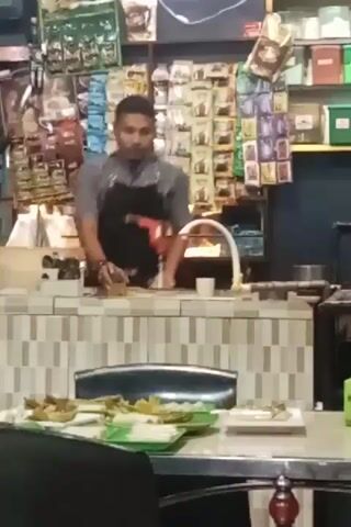 The video shows a man behind a counter in what appears to be a small restaurant or cafe. He is wearing a gray shirt and a black apron and seems to be operating a blender or some other kitchen appliance. Behind him, the wall is covered with various items, including what look like packets of tea or coffee, and shelves with various containers. In front of the counter, there's a table with plates of food. The environment looks somewhat cluttered, suggesting a busy or small establishment.