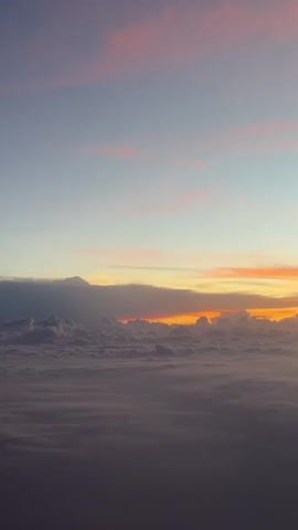 The video presents an aerial view of a sunrise or sunset from above a dense layer of clouds. The sky displays a stunning gradient of colors, with vibrant oranges and yellows near the horizon, transitioning into soft pinks and light blues higher up. The cloud layer below is thick and appears as a sea of grey and white, with some distinct cloud formations visible towards the horizon where the light is most intense. The light subtly brightens and the colors deepen throughout the clip, indicating either an unfolding sunrise or the final moments of a sunset. There are no people or specific objects visible, only the vast sky and cloudscape. While the provided context suggests Sambuor Commune, Cambodia, the visual content itself does not offer specific geographical cues. No text is present in the video.