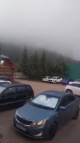 A parking lot is visible with several cars. In the background, there is a wooden building with a green roof and a fence. Trees are visible behind the building. The sky is overcast with fog. The video appears to be taken in the morning or evening.