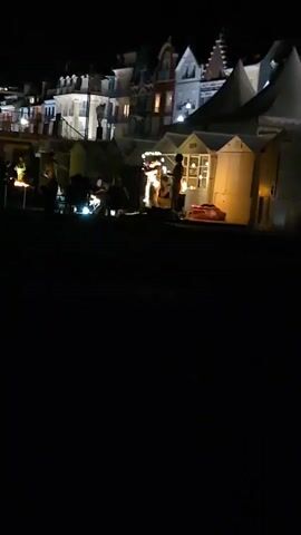 The video shows a nighttime scene with a row of buildings in the background and several small structures in the foreground. People are gathered around some of the foreground structures, and there are lights visible. In the center of the frame, two figures are standing near a well-lit structure. The area in front of the structures is dark.