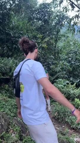 The video captures two individuals trekking through a dense, lush green jungle or forest environment in Nậm Dịch Commune, Vietnam.

The primary subject is a man with a man bun, viewed from behind for most of the clip. He is wearing a white t-shirt with text visible on the back that reads "MAO SAA TREKKING AND HOMESTAY," followed by a phone number and "maosaatrekking.com," accompanied by a logo depicting a person carrying a backpack. He also wears a black fanny pack or crossbody bag. Initially, he moves his arms in a playful or stretching manner before turning and walking forward on an uneven, natural path.

Following the man is a woman, partially visible. She is wearing a black top and a black backpack, and is holding a red jacket. The surroundings are characterized by thick vegetation, including numerous trees, bamboo, and various types of undergrowth, typical of a tropical forest. The lighting suggests it is daytime, with diffused light possibly due to an overcast sky or dense tree canopy. The overall activity is an outdoor trek, with the individuals navigating through the natural terrain.