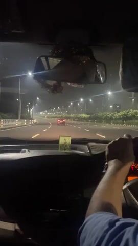 The video shows the view from inside a car at night, looking forward through the windshield and past the dashboard. The driver's right arm and hand are visible on the steering wheel, which is turned slightly to the right. In the distance, a highway stretches ahead, illuminated by streetlights and the headlights of other vehicles. Two red taillights are visible on a car ahead, and numerous streetlights line both sides of the highway. The sky appears dark and possibly hazy, and there is a rearview mirror hanging from the windshield, with some decorative items attached to it. A small religious figurine is placed on the dashboard. The car appears to be moving at a moderate speed.