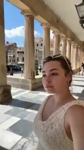 A young woman is standing in an arcade composed of columns. She is wearing a white lace dress and sunglasses are perched on her head. The flooring of the arcade is a gray and white checkerboard pattern. Beyond the arcade is a courtyard or square, with buildings visible in the background under a clear blue sky. The woman glances at the camera, offering a slight smile.