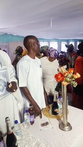 Here's a description of the video:

The video shows a gathering of people indoors, likely at a wedding or celebratory event. Many are dressed in white, which is a common color for such occasions in some cultures. There are tables set up with flowers, drinks, and decorative items. People are milling around and talking. The environment appears festive and social. There are a few people in traditional Nigerian dress as well. The ceiling is decorated with fabric, giving the space a tent-like feel.