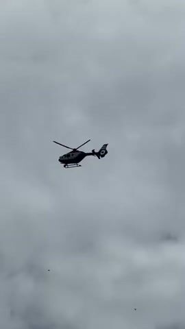 A helicopter flies across a cloudy sky. There is a small object falling from the helicopter. The helicopter is black and white with a red stripe. It is in the center of the frame and moving from left to right. The sky is overcast with gray clouds. The object falling is small and dark.