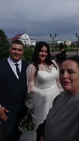 A group of people are outdoors, possibly at a wedding. Three people are prominently featured in the foreground, with a couple in the center, and a woman to the right. The couple appears to be a bride and groom. The bride is wearing a white wedding dress with lace sleeves. The groom is in a dark suit with a vest and tie. The woman on the right is wearing a grey top. In the background, there are buildings, streetlights, trees, and a cloudy sky. Some people are visible in the background as well, including a person who appears to be taking a photo. The video appears to be a selfie-style recording as the camera angle is dynamic.