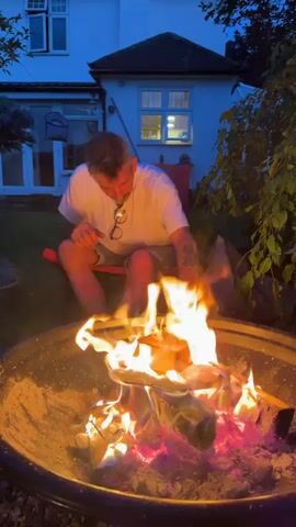 A man is sitting in a red folding chair in a backyard at dusk, opening a bottle of wine. In front of him is a large, round, metal fire pit with a fire burning brightly inside. The fire consists of logs and what appears to be some kind of flammable material on top, creating high flames. The house behind him is a two-story building with white siding and visible windows, one of which is lit. To the right of the man is a tree with green leaves. The sky is a deep blue, suggesting it is early evening. The man is wearing a white t-shirt, shorts, and glasses hanging from his shirt. He appears to be focused on opening the wine bottle.