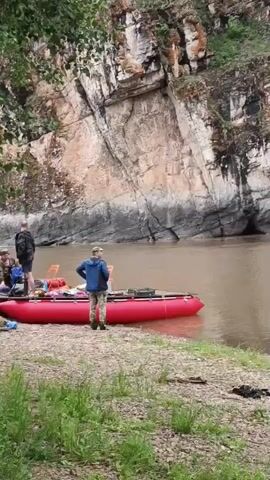 The video shows a scenic riverbank with people preparing for or engaged in water activities. In the foreground, two individuals stand near a large, red inflatable raft on a rocky, grassy shore. The raft is laden with camping gear, including backpacks and orange containers. Further down the river, another inflatable raft is visible, this one crowded with several people, appearing to be a tour group or a rafting expedition. One person stands at the back of this raft, possibly guiding it. The river itself is a murky brown color, and it flows past a dramatic, rocky cliff face covered in sparse vegetation. The overall impression is of an outdoor adventure in a natural, somewhat wild setting.
