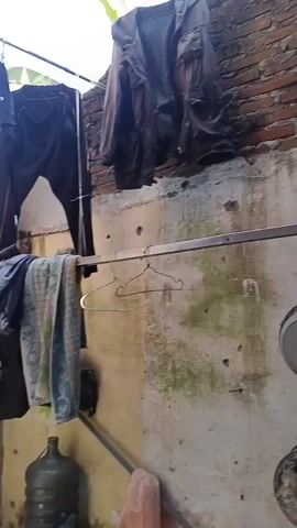 The video shows a clothesline with various items of clothing hanging on it, including shirts, pants, and a jacket. There are also several empty hangers and some buckets on the ground. The scene appears to be an outdoor area, possibly a backyard, with a brick wall in the background. The lighting suggests it is daytime. The camera pans slowly from left to right, showing the clothesline and the surrounding area.
