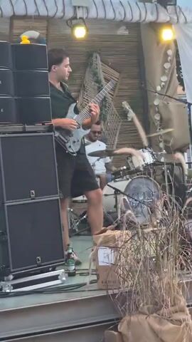 A band is performing live on an outdoor stage. In the foreground, a man in a dark green short-sleeved shirt and black shorts plays a silver electric guitar. He is standing on the left side of the stage. To his right, and slightly behind him, a man in a white t-shirt and blue shorts plays the drums. He is wearing sunglasses. The stage is decorated with a beach theme, with netting, driftwood, and seashells visible in the background. There are large speaker cabinets on the left side of the stage. A bottle of beer is on the floor near the guitar player. Tall, dried grass is in the foreground, partially obscuring the view of the stage. The lighting appears to be natural daylight mixed with stage lights.