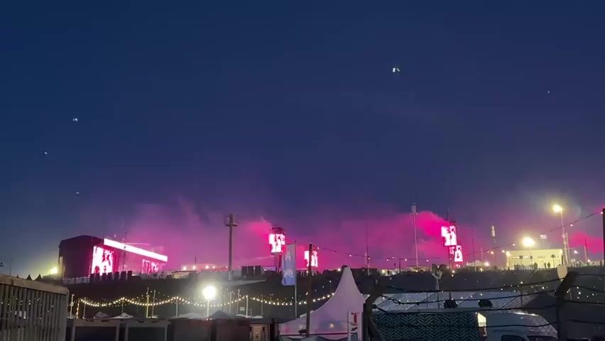 The video shows an outdoor festival or concert venue at night. The sky is dark blue. In the background, multiple large structures, resembling stages or towers, emit plumes of colored smoke that change from blue to yellow, then to pink and red, and back to blue. Some of these structures have large illuminated screens. A crowd of people is visible on a raised area in the distance. The midground features a gently sloping terrain with string lights and several tall light poles. In the foreground, a wire fence, a pink tent, and a white van are visible. The overall scene is brightly lit with various lights.