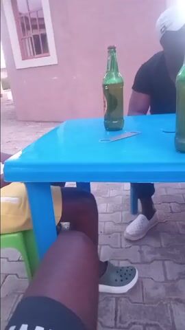 The video shows three people sitting around a light-blue plastic table. Two green beer bottles and a bottle opener are on the table.  One man wears a white top and yellow shorts and sits on a green plastic chair. Another man wears a black top and black pants and is seen opening another beer bottle. The person filming is also seated at the table, wearing dark shorts and green shoes. They are sitting outdoors, possibly in a courtyard or patio, with a peach-colored building in the background. The sky appears to be overcast. There is some conversation taking place.