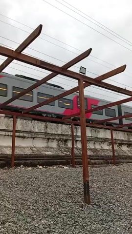 A train is moving across the screen from right to left. The train is light grey with a thick horizontal red stripe. The train passes behind a series of wooden post and beam structures, and behind a concrete retaining wall that runs parallel to the tracks.  The foreground is gravel. The sky is overcast. A man wearing a hat and sunglasses walks into the scene on the bottom right.