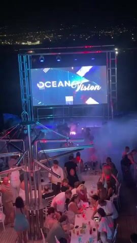 The video depicts a night scene at an outdoor venue, likely a rooftop or a boat, illuminated by colorful lights and a large LED screen displaying "OCEAN Vision". Several tables are occupied by groups of people, with drinks and bottles visible on them. Some individuals are standing and dancing, while others are seated. Smoke or fog machines are in operation, adding to the atmosphere. The background shows a distant city skyline at night. The overall impression is that of a lively party or club environment.