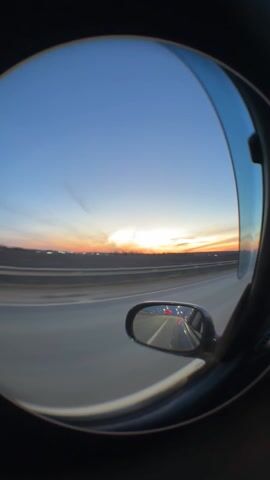 A fisheye lens captures a sunset view from a car traveling on a highway. The sky transitions from blue to orange near the horizon. The highway is visible with a barrier along the side, and cars are seen in the car's side mirror. The footage is captured from inside the car, framing the sunset view.