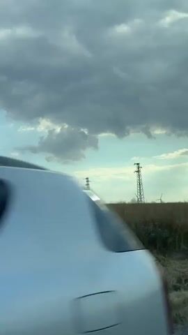 A video captured from a moving vehicle shows a rural landscape under a cloudy sky. The foreground is dominated by dry grass and vegetation, partially obscuring a metal guardrail. In the mid-ground, a field of tall, dry grass or crops stretches across the frame. In the background, two tall transmission towers with visible wires stand out against the sky. Further in the distance, several wind turbines are visible on the horizon. The sky is a mix of light blue and dark, heavy clouds, suggesting overcast or approaching weather. The overall impression is one of an open, agricultural area with infrastructure for power generation.