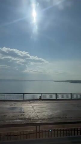 The video captures a wide shot of a body of water viewed from a high vantage point, possibly a bridge. The sun is bright and high in the sky, casting a lens flare and creating beams of light that shine down through scattered clouds. In the foreground, there is a paved surface with a metal railing. The railing runs horizontally across the middle of the frame, separating the paved area from the body of water. Beyond the railing, the water stretches to the horizon, meeting a hazy sky. The water appears calm, with a few distant landmasses visible on the far shore. The overall impression is one of a bright, sunny day overlooking a vast expanse of water.