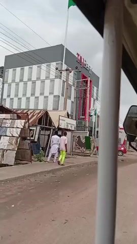 The video shows a street scene with a modern bank building in the background. The bank has "TAJ Bank" written in large letters on its facade. There are flags in front of the bank, one of which appears to be the Nigerian flag. A red car is parked in front of the bank. In the foreground, two men are walking on the street. One man is wearing a white outfit, and the other is wearing a pink shirt and neon green pants. There are also some stalls or shops with corrugated metal roofs along the side of the street. The sky is overcast. The video is shot from the perspective of someone in a vehicle.