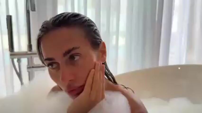 A woman is in a bathtub full of bubbles, holding a black smartphone and looking at the screen. Her hair is wet and slicked back. She is leaning her face on her hand. A chrome faucet is visible next to her. Behind her is a large window with sheer white curtains.