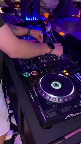 This media file shows a close-up, high-angle shot of a person's hands operating a DJ setup in a dimly lit venue. The DJ is wearing a watch and a ring on their left hand, and has tattoos on their arm. They are wearing white shorts and white sneakers. The DJ equipment includes a Pioneer DJ mixer and what appear to be Pioneer CDJ-2000 Nexus CD players. The equipment is lit with colorful LEDs, primarily blue and orange, creating a vibrant atmosphere.

In the background, the setting appears to be a bar or club, with tables and chairs visible, and other patrons blurred in the distance. There is a metallic structure with blue lighting behind the DJ. The overall mood is energetic and focused, suggesting a live performance.

The visible text on the equipment includes "Pioneer," "CDJ-2000 Nexus," and what appears to be a cable label that reads "PROFESSIONAL CABLE." The time of day is likely evening or night due to the lighting and the nature of the venue. There are no discernible weather cues as the scene is indoors. The location context provided is Tiraspol, Moldova.
