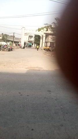 A horizontal shot shows a road with a building on the left. The road is paved and appears to be a street. The building is white with a large archway. There are people and motorcycles near the building. The sky is clear and the sun is shining. The video is shaky and the angle is tilted.