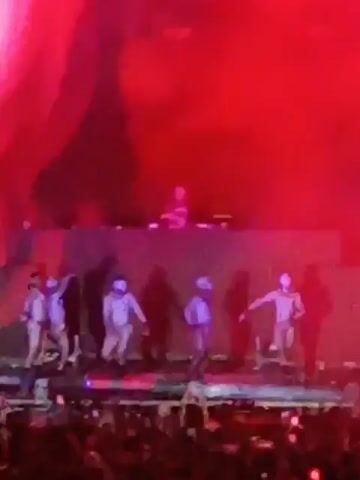 The video shows a music concert with a large crowd of people waving their hands in the air. On a stage above them, a DJ stands at a control panel, and several performers in white outfits are dancing. Red lights are flashing on the stage. The backdrop behind the performers is a large screen displaying abstract shapes. The overall atmosphere is energetic and chaotic.