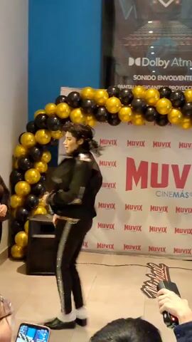 The video shows a Michael Jackson impersonator performing at an event. The impersonator is wearing a black jacket with silver accents, black pants with a silver stripe, white socks, and black shoes. They have long, dark, curly hair and are performing a dance routine.

The scene is set in a cinema lobby, with a blue wall and a large banner advertising "MUVIX CINEMAS" in red letters. A balloon arch made of black and gold balloons frames the performance area. In the background, there are posters for movies, including one with the "Dolby Atmos" logo. Another poster on the right features Michael Jackson with the text "MICHAEL JACKSON" and a QR code.

The event appears to be taking place indoors, likely during the day given the lighting. The atmosphere seems festive, with the balloon arch and the presence of an audience. The impersonator's movements suggest they are engaged in a lively performance. The presence of multiple camera phones suggests that the audience is actively capturing the event.