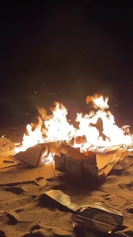 A campfire burns brightly at night on a sandy beach. Cardboard boxes are the primary fuel, visibly breaking down as they ignite. Flames reach upwards, casting an orange glow on the surrounding sand. The background is dark, indicating the nighttime setting. The camera is positioned at a low angle, focusing on the fire.