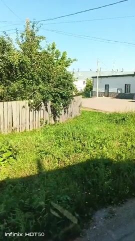 The video shows a street scene in what appears to be a rural or suburban area on a sunny day. A wooden fence is visible on the left side of the frame, with lush green grass and some wildflowers growing alongside it. A paved road runs through the center of the frame, leading towards a white building with a green trim and a dark roof. There are several windows and doors on the building. A silver sedan is parked in front of the building, and a person is seen in the driver's seat, though their features are not clearly discernible. Power lines are strung across the top of the frame. The overall impression is of a quiet, ordinary day in a small town or village.