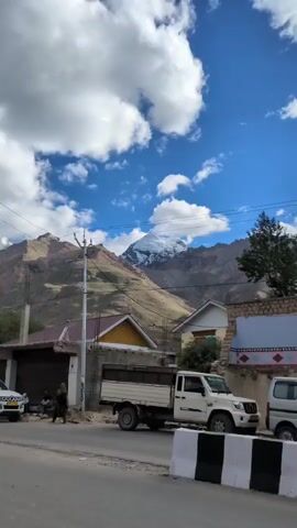 The video pans across a mountainous landscape under a bright blue sky with scattered white clouds. In the foreground, there are several buildings, a white pickup truck, a flatbed truck, and a white van parked along a street. A utility pole with wires stretches across the frame. In the background, large mountains rise, with one prominent peak covered in snow. The overall scene suggests a rural or small-town setting in a mountainous region.