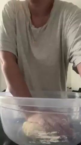 A woman stands in what appears to be a kitchen, kneading dough in a large, clear plastic bowl. She wears a light grey T-shirt and a gold necklace. Her hands are covered in flour. In the background is a modern chandelier and the reflection of a television screen. There is music playing in the background.