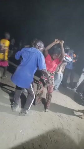 The video shows a group of people dancing outdoors at night. A man in a blue shirt and black pants is dancing with a woman wearing a red shirt and patterned pants. She has her hands raised in the air. Another man, wearing a black tank top and shorts, stands to their left and also appears to be dancing. Other people are visible in the background, some standing and watching. The ground appears sandy. The lighting is dim, suggesting a nighttime setting. Music is audible in the background.