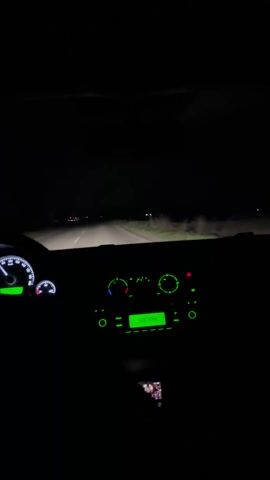 The video shows the interior of a car driving at night on a rural road. The dashboard is illuminated in green. The radio display shows the frequency 106.5 MHz. The car's headlights illuminate the road ahead. Someone is briefly seen looking at a phone. The sound of music plays in the background.