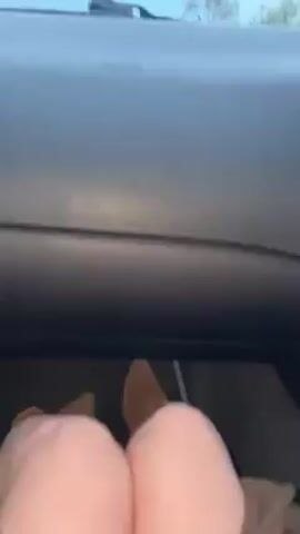 The video shows a car driving on a road along the coast. The camera is inside the car, looking out over the dashboard. The driver is wearing brown suede boots. The road has a concrete barrier with black and white striped directional arrows. There is a body of water visible in the distance, and some landmass on the horizon. The sky is cloudy and the light is dim.  The audio suggests music is playing in the car.