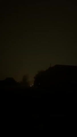 The video is dark and grainy, with the majority of the frame obscured by shadow. Silhouetted trees and buildings are visible against a hazy, dimly lit sky, suggesting nighttime or heavy cloud cover. There are faint lights in the distance, possibly from streetlights or windows. No people or specific actions are discernible due to the poor lighting and low resolution.