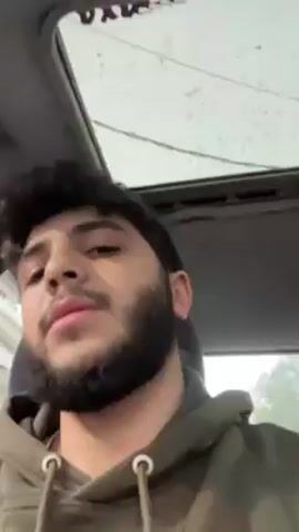 A medium shot from inside a car shows a young man with dark curly hair and a full beard wearing a green hoodie. He is looking directly at the camera and talking. Above him is the car's sunroof, which appears to have raindrops on it. Outside the car window, there is a glimpse of green trees. The overall lighting in the car is soft, indicating overcast weather.