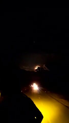 The video shows a night scene on a road. The camera is in motion, likely from a vehicle. The road stretches out ahead, illuminated by the headlights of a vehicle, casting a yellow glow on the road surface. The sky above is dark, with the moon partially obscured by clouds, creating a glowing effect around the edges of the clouds. A set of two bright objects, possibly stars or lights, are visible higher in the sky. A dark silhouette, likely a person's shoulder or back, is in the foreground of the shot. The overall impression is a night journey on a rural road, with the moon and passing vehicle lights as the main sources of illumination.