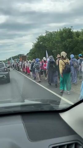 The video shows a procession of people walking along the side of a road in a rural or suburban area. The group appears to be a mix of men, women, and possibly some older children, dressed in a variety of casual clothing. Many of the women are wearing headscarves or hats, and some are wearing long skirts or dresses. The people are walking in a line, and some are carrying backpacks. There are flags visible among the group, suggesting a possible organized event or protest.

The scene is set on a paved road with a white line marking the shoulder. On the right side of the road, there is a line of trees and vegetation. On the left side, there are other vehicles, including a dark grey SUV, and what appear to be buildings or structures in the distance. The sky is overcast with clouds, indicating a possibly cool or damp day. The lighting suggests it is daytime, likely late morning or afternoon.

The overall mood appears calm, with people walking steadily. There are no overt signs of distress or agitation. The presence of multiple flags and the organized formation of the group suggest a planned event, possibly a pilgrimage, protest, or cultural gathering. The visual cues, such as the clothing and headscarves, along with the location context of Sadovodov, Russia, may hint at a religious or cultural event specific to the region. There is some text visible on the license plate of the grey SUV, which reads "B024BY 122". Other text on the flags is not clearly legible.