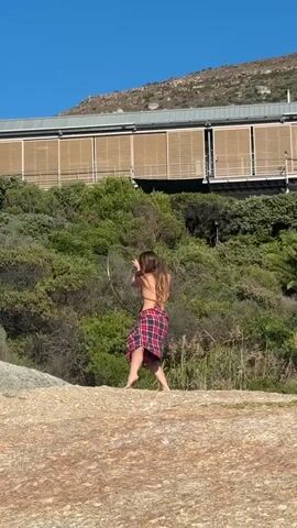A young woman with long brown hair is seen outdoors on a rocky, sandy surface. She is wearing a red and black plaid skirt or wrap and what appears to be a dark bikini top. She is barefoot and actively engaged in hula hoop tricks, twirling a golden hoop around her body, throwing it into the air, and catching it. Behind her, there is dense green foliage consisting of bushes and trees. Further in the background, a modern building with numerous horizontal, light-colored slatted panels is visible, seemingly built into a hillside. A large mountain rises in the distance under a clear, bright blue sky. The scene is well-lit, indicating a sunny day. A male voice speaking in Russian can be heard in the background.