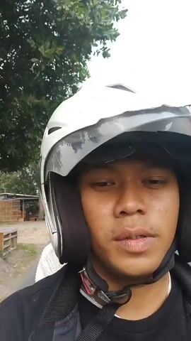 A person wearing a white helmet and black jacket is riding on a motorcycle. They are filming themselves with a camera. Behind them, another person wearing a helmet and a white sweater is visible. The background shows trees and a road, suggesting an outdoor setting.