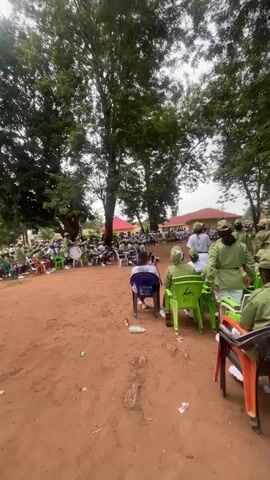 The video shows a large group of people, mostly young adults, gathered outdoors under trees. Many of the individuals are wearing matching green uniforms, consisting of shirts and hats, and some are also wearing white t-shirts and skirts or shorts. They are seated in plastic chairs and standing in a dirt-filled area. Some people are looking towards a central point, suggesting an event or gathering is taking place. The environment appears to be a school or camp setting with buildings visible in the background. The lighting suggests it is daytime, possibly overcast. There is a drum visible in the background.