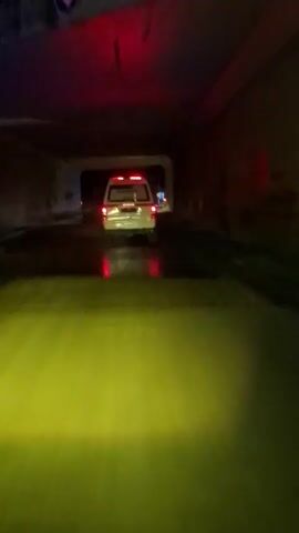 The video shows a white van driving through a dark tunnel. The tunnel walls are concrete and dimly lit by the van's headlights and red lights from the rear of the van. As the van exits the tunnel, it continues to drive on a road at night. There are streetlights and other vehicles on the road. The road is wet, and reflections of the lights are visible on the surface.